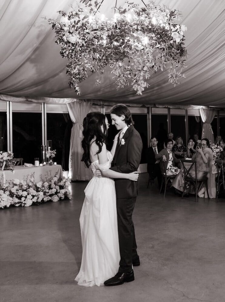A timeless black and white photo of a couple’s first dance under hanging greenery in a tented reception.