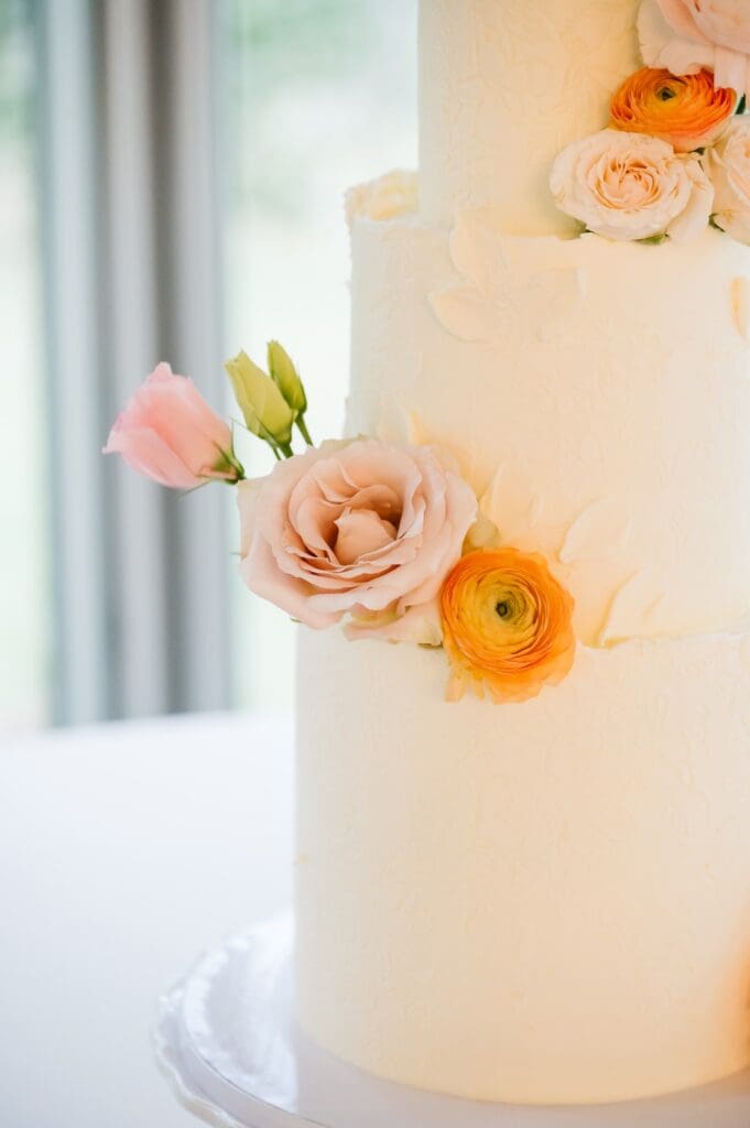 A white tiered wedding cake with delicate fresh floral accents featuring peach ranunculus and garden roses, curated for a romantic celebration at Hummingbird House.