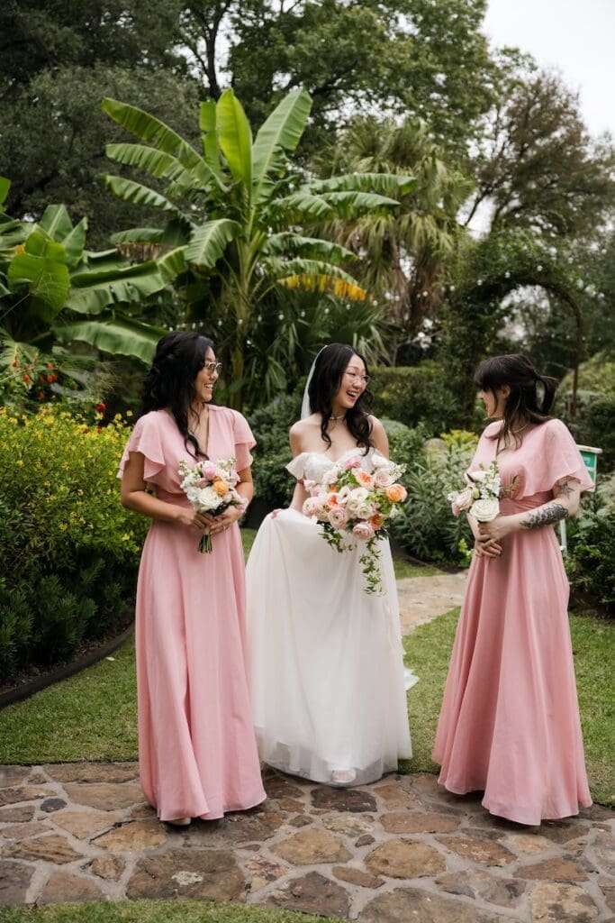 Bride and bridesmaids in elegant pink gowns laughing together in a lush tropical garden setting in Austin.