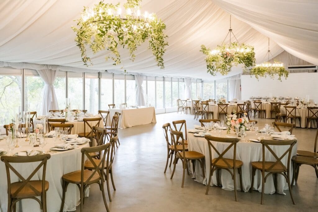 An elegant tented wedding reception featuring chandeliers draped in greenery and fluted glass tabletop details.