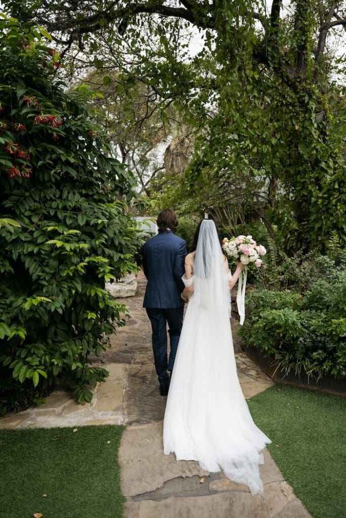 A romantic back-view portrait of a bride and groom walking along the signature stone path at a Hummingbird House garden wedding.